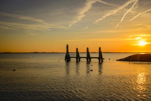 sunset in the northsea with lines from planes in the sky and wooden structure in the water