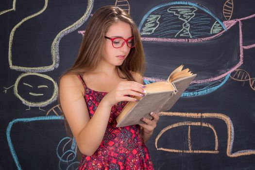 Young student girl in red dress and red glasses reads a book in front of school blackboard
