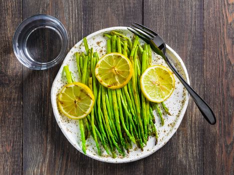 Grilled asparagus and slice of lemon in craft trendy plate on dark brown wooden table. Top view or flat lay.