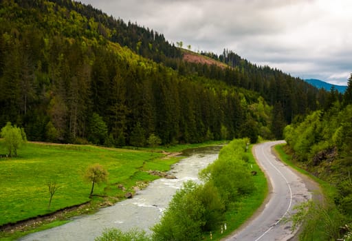 road along the river near the forest on hillside. lovely springtime landscape in mountains on a cloudy day. view from above