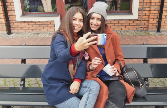 Two young women make selfie sitting on a bench outdoors
