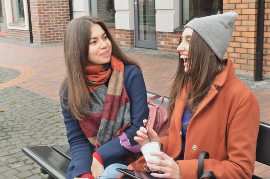 Two attractive girls are sitting on a bench in the street near the trees and buildings, holding coffee and a plate in their hands, laughing