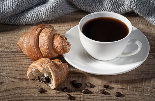 Black coffee with croissants. Coffee grains on a wooden table. Knitted woolen scarf in the background.
