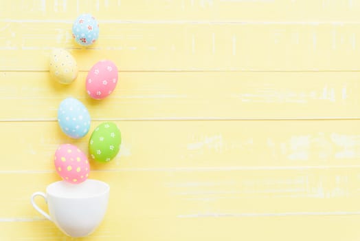 Happy easter! Row colorful Easter eggs spread out from white cup on bright yellow wooden background.