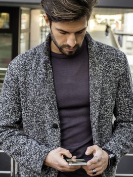 Handsome trendy man wearing cardingan standing and typing on cell phone, outdoor in city setting in day shot