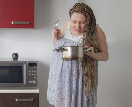 Hungry pregnant woman is holding a saucepan and a spoon, sticking her tounge out.