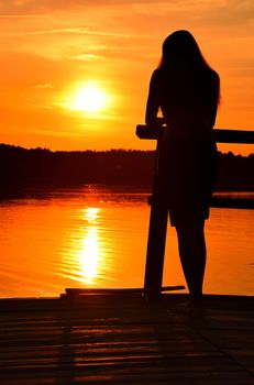 Sunset silhouette of one young woman with long hair standing on lake or river wooden pier and looking away, rear view