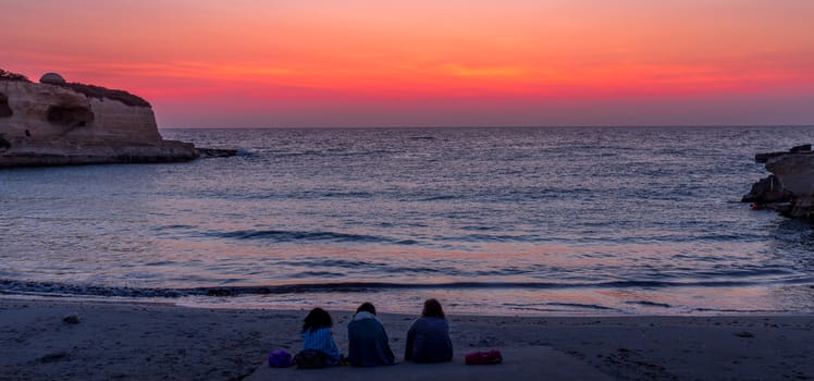Three women waiting for the sunrise in front of the sea (Puglia region, South of Italy). Concept of frienship, travel and adventure.