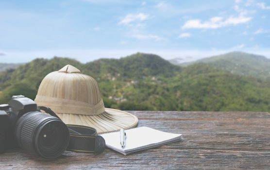 Camera with cap, notebook and pen on wooden table with mountain and sky background, Travel concept. Travel Accessories.