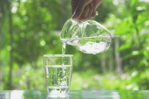 Female hand pouring water from pitcher to glass on nature background