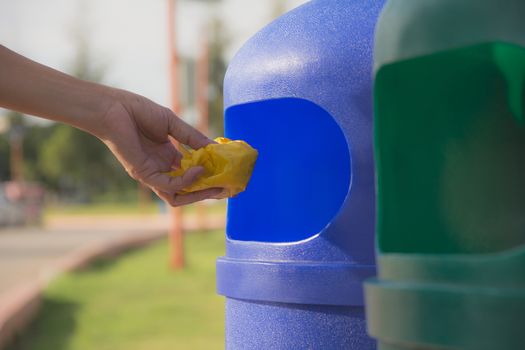 Female hand throwing crumpled yellow plastic into blue plastic trashcan.