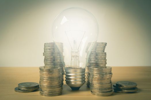 Energy saving light bulb and surrounding by stacks of coins on wooden table.