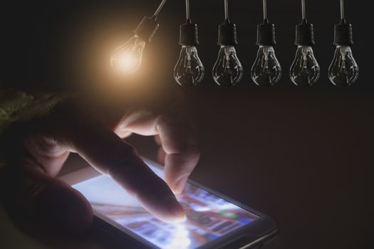Hanging light bulbs with glowing one with hand touching screen of smart phone on dark background. Idea and creativity concept with light bulbs.