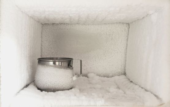 Stainless steel drinking water glass in freezer of a refrigerator. Ice buildup inside of a freezer walls.