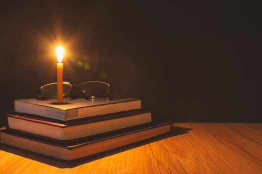 Close-up of burning candle with pile of old book on wooden table. Education concept.