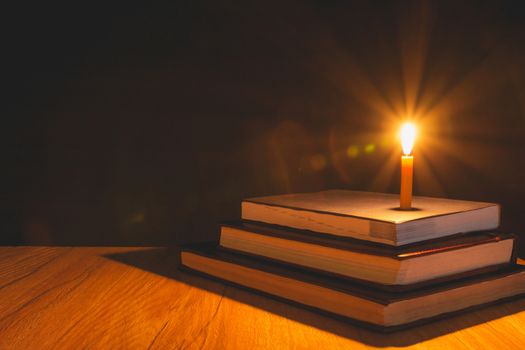 Close-up of burning candle with pile of old book on wooden table. Education concept.