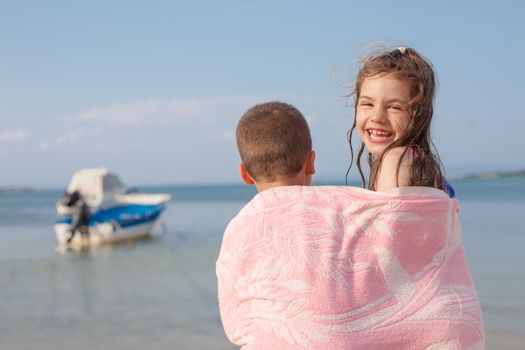 A girl looking to camera smiling and a boy with his back to the viewer, both sharing a towel