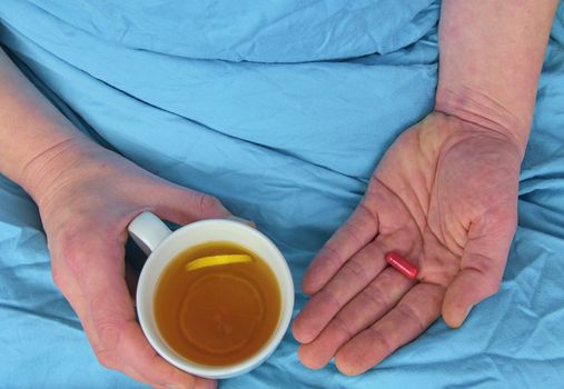 Influenza season. Hands of the sick man holding pill and cup of the hot tea in the bed. Close-up of ill caucasian man at home.