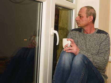 Depressed middle age man sitting near window. Sad man drinking coffee.