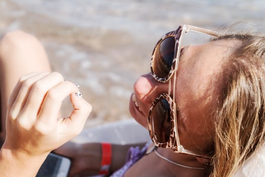 A young tanned woman looks at her nail-manicure and laughs at the smartphone through sunglasses on a deckchair. The concept of a relaxed lifestyle.