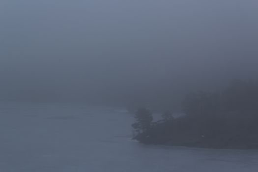 Frozen lake surrounded by small mountains with pine and fir trees on a misty, moody, day. Pine and fir trees on a mountain leading down to a frozen lake on a misty day. Nackareservatet - nature reserve in Sweden