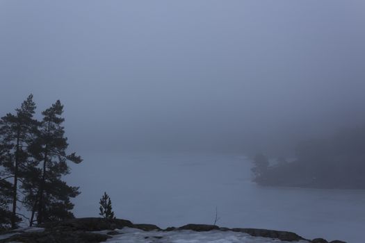 Frozen lake surrounded by mountains with pine and fir trees on a misty, moody, day. Nackareservatet - nature reserve in Sweden