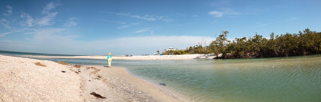 Older woman stands on the white sand beach in front of aqua blue water of Clam Pass in Naples, Florida in the morning.