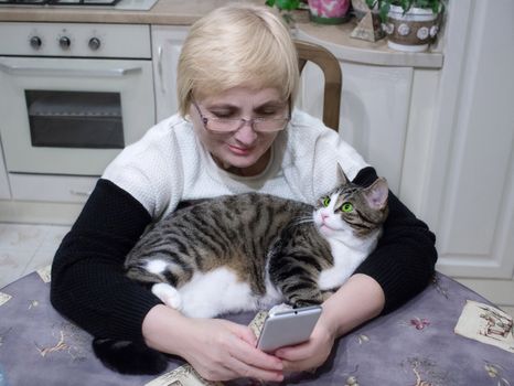 Elderly woman sends sms on mobile phone sitting at kitchen table with pet cat