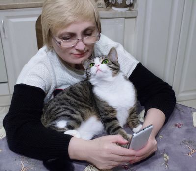 Elderly woman sends sms on mobile phone sitting at kitchen table with pet cat