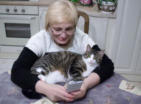 Elderly woman sends sms on mobile phone sitting at kitchen table with pet cat