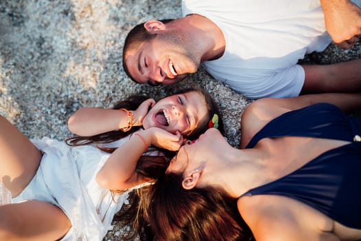 Young happy family walking along the seashore. Little girl with her parents lying on the sand and smiling, parents want to kiss their girl on the cheek.