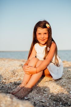 Little beautiful girl walking along the seashore at the day time. She is sitting on the sand, wearing white dress, long brown hair.