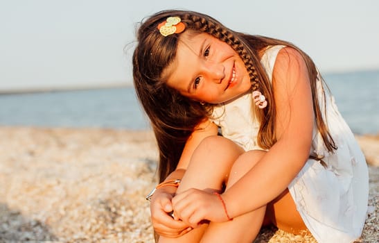 Little beautiful girl walking along the seashore at the day time. She is sitting on the sand, wearing white dress, long brown hair.