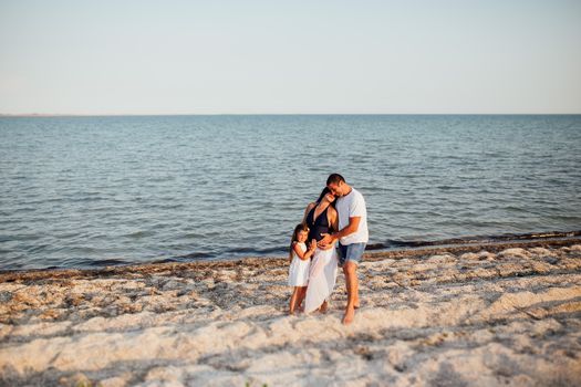 Young happy family walking along the seashore. Little girl with her parents walking on the beach, they standing, holding each other and embracing mother's tummy, she is pregnant.