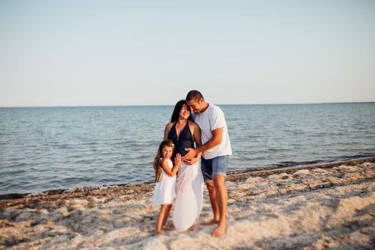 Young happy family walking along the seashore. Little girl with her parents walking on the beach, they standing, holding each other and embracing mother's tummy, she is pregnant.