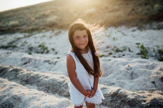 Little beautiful girl walking along the seashore at the day time. She is wearing white dress, long brown hair.