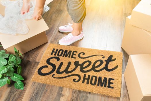 Woman in Pink Shoes and Sweats Unpacking Near Home Sweet Home Welcome Mat, Moving Boxes and Plant.