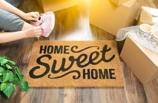 Woman Wearing Sweats and Untying Pink Shoes Near Home Sweet Home Welcome Mat, Moving Boxes and Plant.