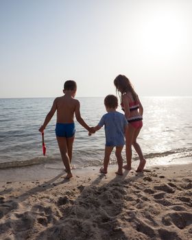 Three kids on the shore of a sea or ocean. Two are leading smaller one to the water.