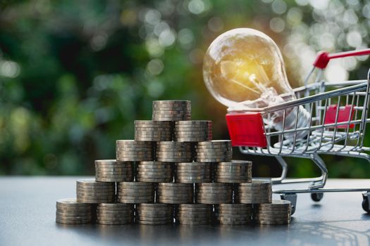 Energy saving light bulb with shopping cart and stacks of coins on nature background. Saving, accounting and financial concept.