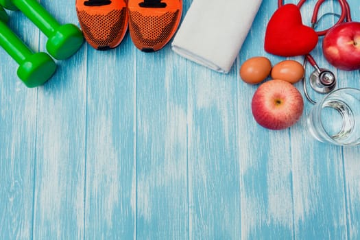 Top view of sportswear, sports equipment, dumbbells and stethoscope on blue and wooden table.