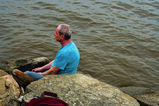 Midle aged man sitting on the shore of the lake. Solitude mature man sitting on the bank. Concept of loneliness. 