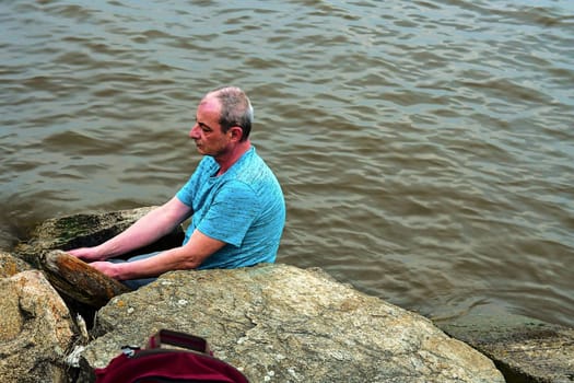 Midle aged man sitting on the shore of the lake. Solitude mature man sitting on the bank. Concept of loneliness. 