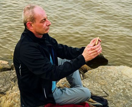 Midle aged man sitting on the shore of the lake. Solitude mature man taking selfie picture  on the bank. Concept of loneliness. 