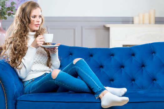 girl with a cup of coffee in a warm sweater and jeans on the couch in the living room