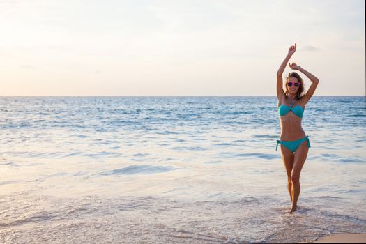 Young, slim, tanned caucasian woman in bikini and sunglasses standing and dancing in the sea