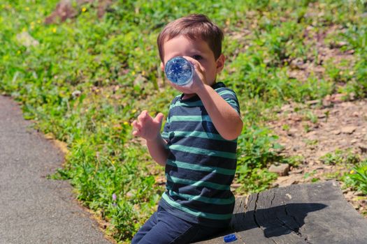 Thirsty little boy sitting on a wooden bench and drinking water.