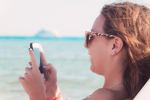 A young tanned woman looks and laughs into a smartphone through sunglasses on a lounger. The concept of a lifestyle is always on the Internet.