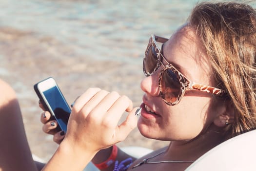 A young tanned woman looks and laughs into a smartphone through sunglasses on a lounger. The concept of a lifestyle is always on the Internet.
