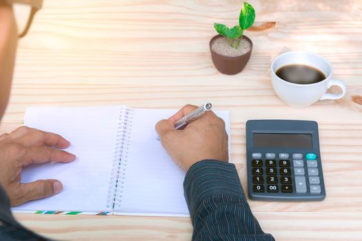 Person working and writing on notebook with calculator on wooden table background.